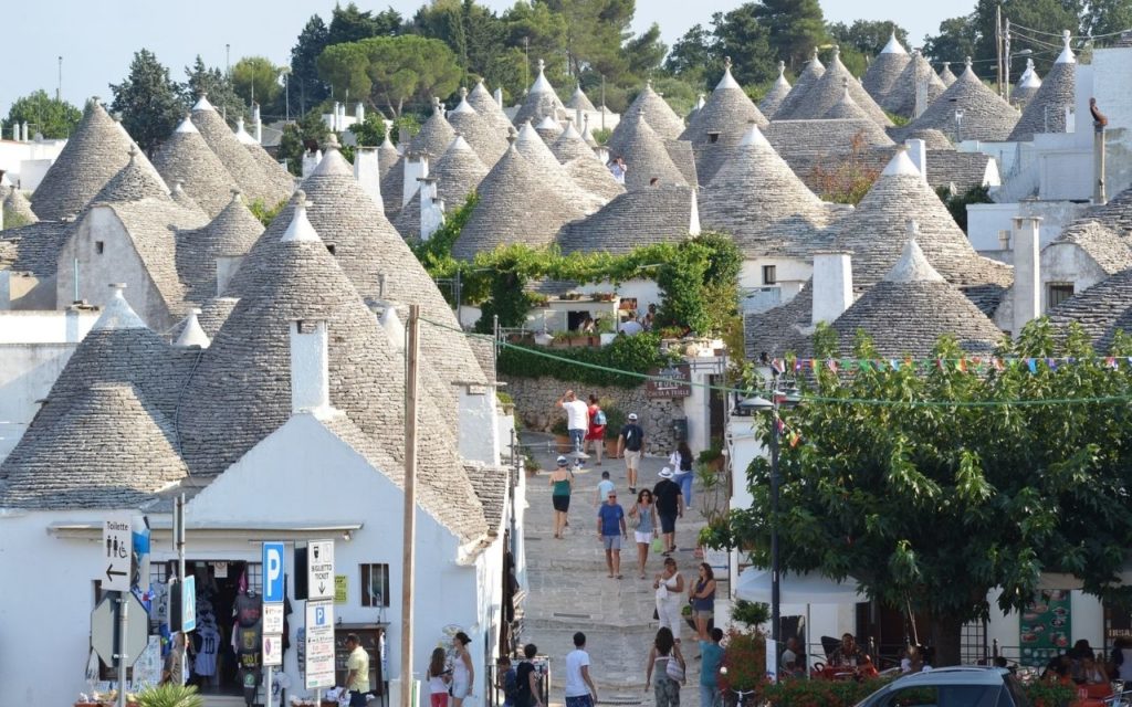 a group of people walking down a street with a group of conical roofs with Alberobello in the background