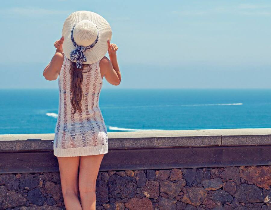 a woman wearing a white dress and hat standing by a stone wall