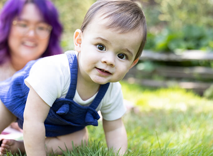 a baby crawling on grass with a woman in the background