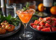 a glass of orange liquid with a rosemary sprig next to a plate of fruit