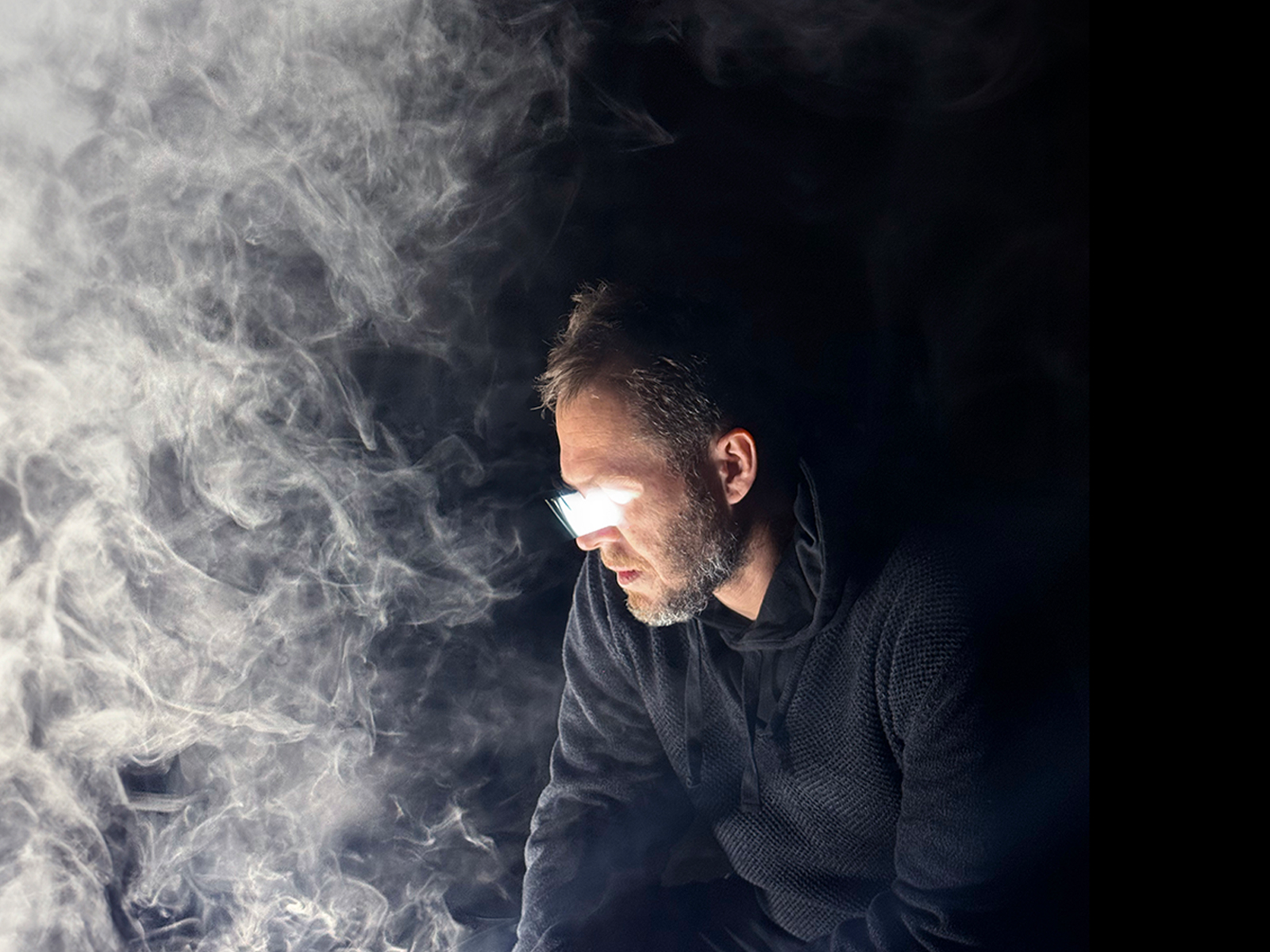 a man with glasses and a beard sitting in a dark room with smoke
