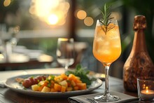 a glass of orange liquid with a rosemary sprig on top of a plate of food