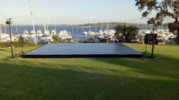 a large black rectangular platform on grass with boats in the background