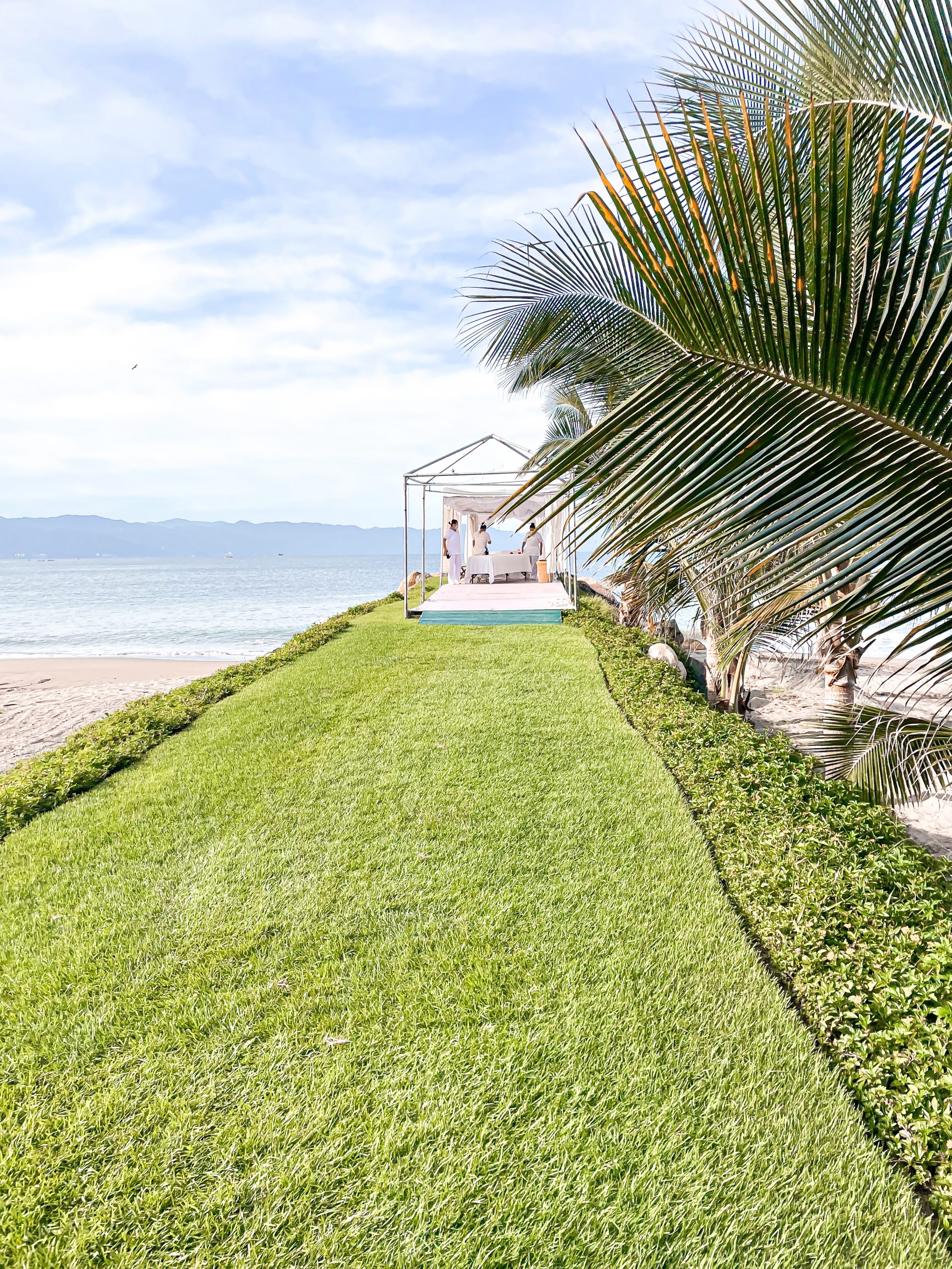 a lawn with a white structure and palm trees on the beach