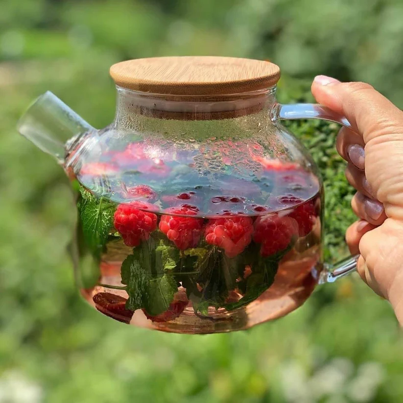 a hand holding a glass teapot with a raspberry and mint