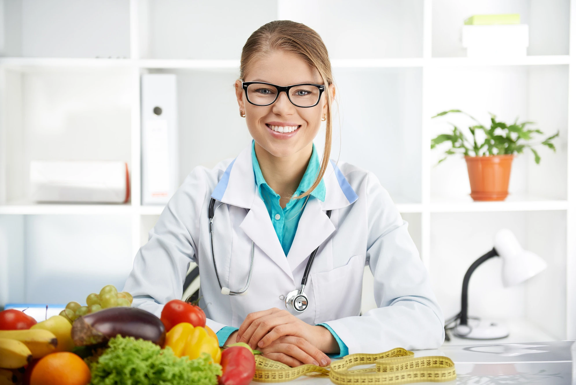 a woman wearing glasses and a stethoscope sitting at a table with a pile of vegetables