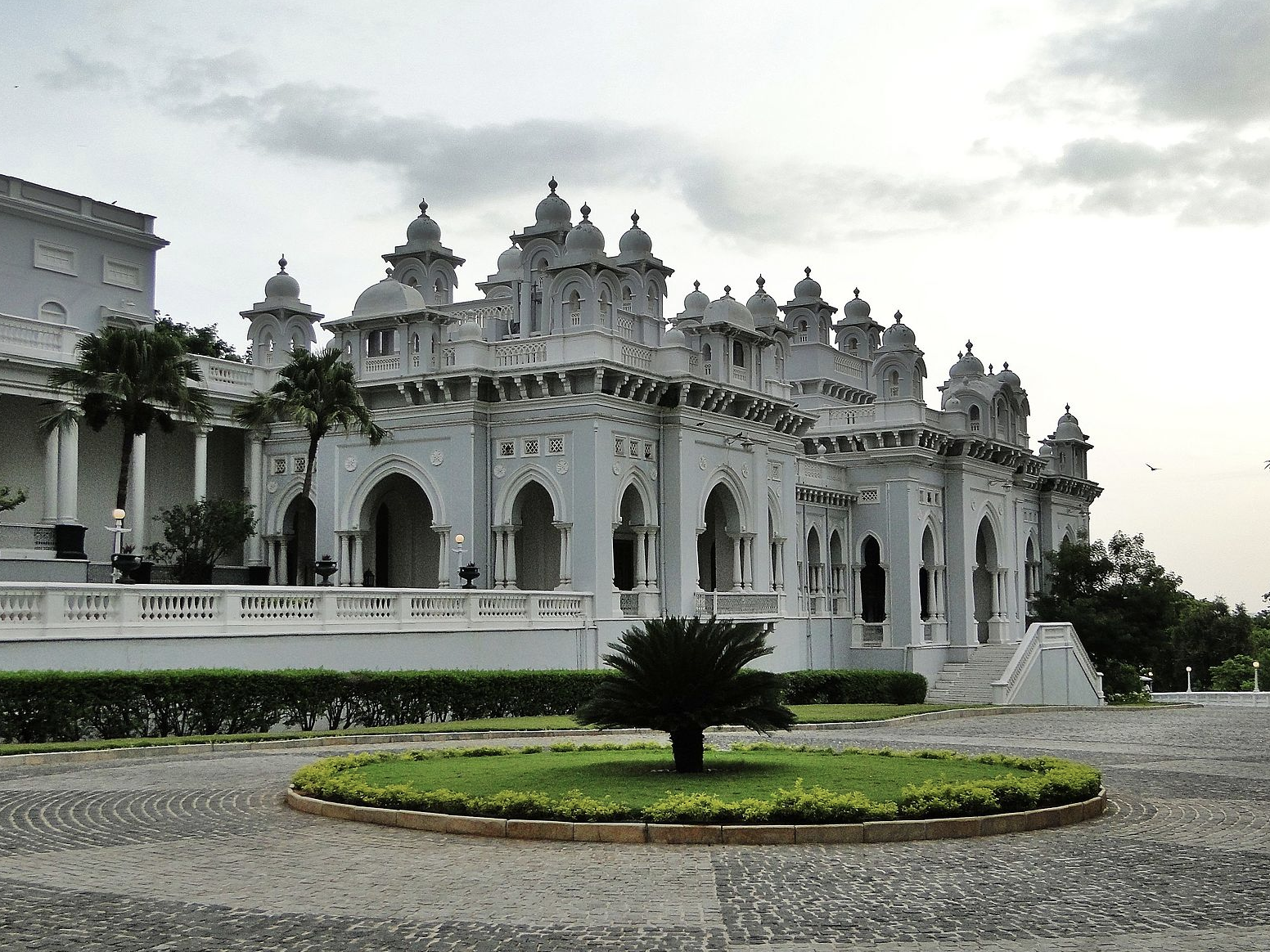 a white building with a circular lawn and a tree in front with Chowmahalla Palace in the background