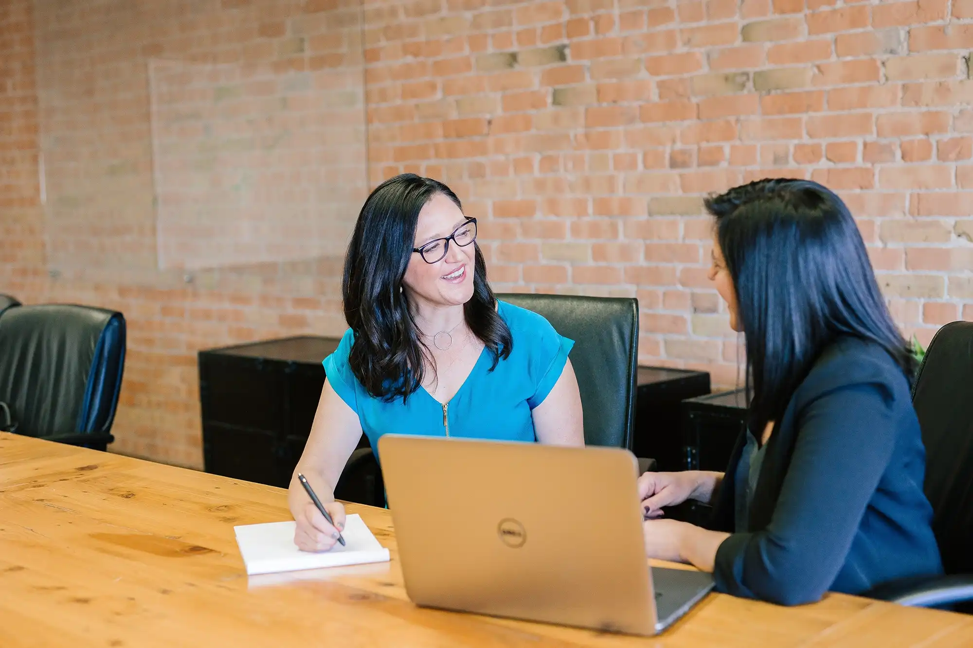 two women sitting at a table with a laptop and a notepad