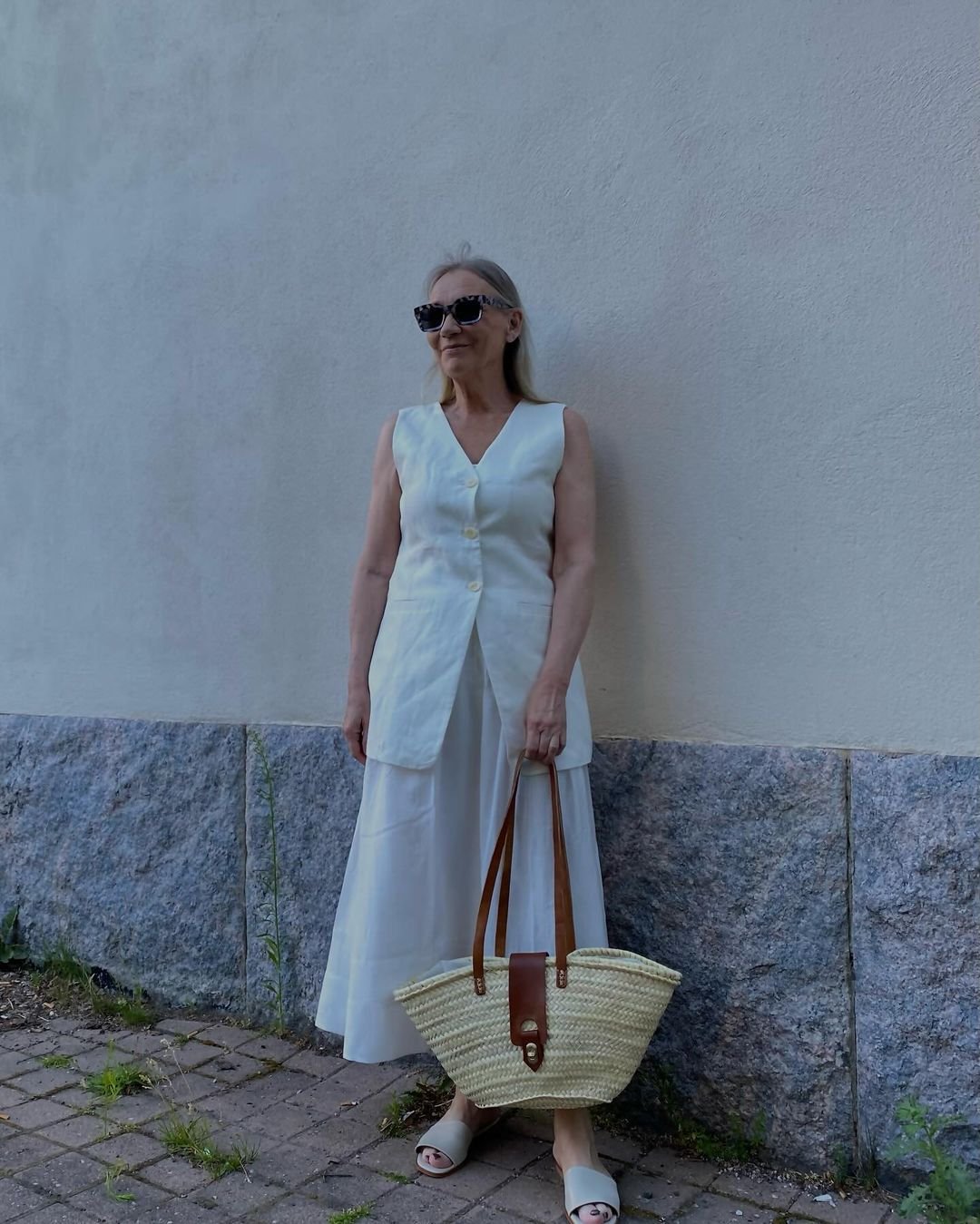 a woman in a white dress and sunglasses standing by a wall