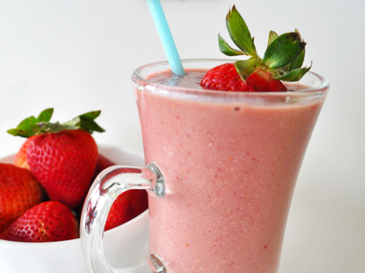 a glass of pink smoothie with straw and straw in front of a bowl of strawberries