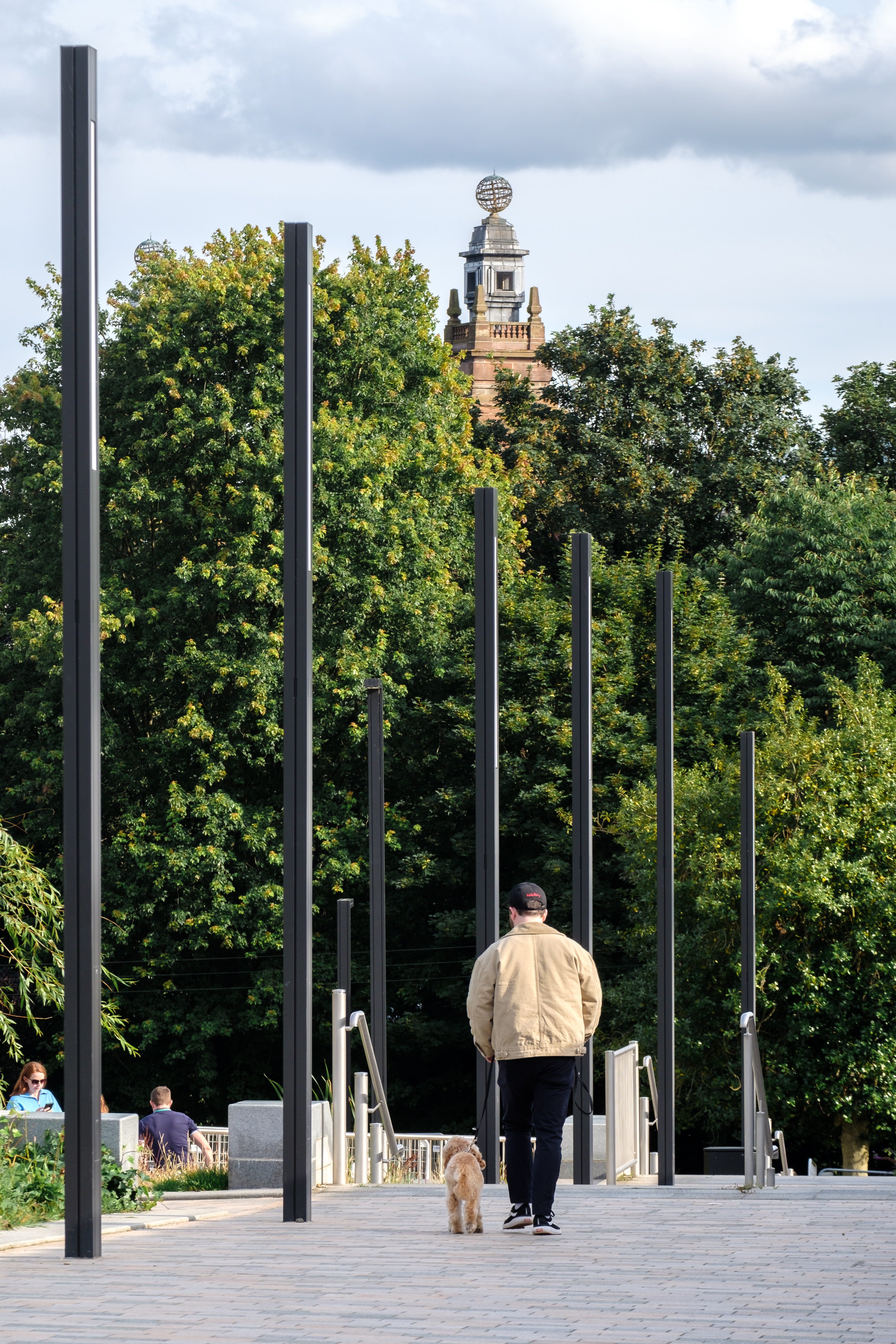 a man walking down a walkway with tall poles and a tower in the background