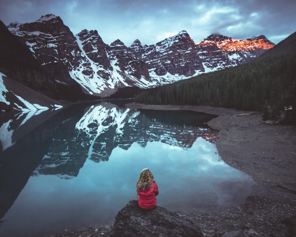 a woman sitting on a rock by a lake with snow covered mountains