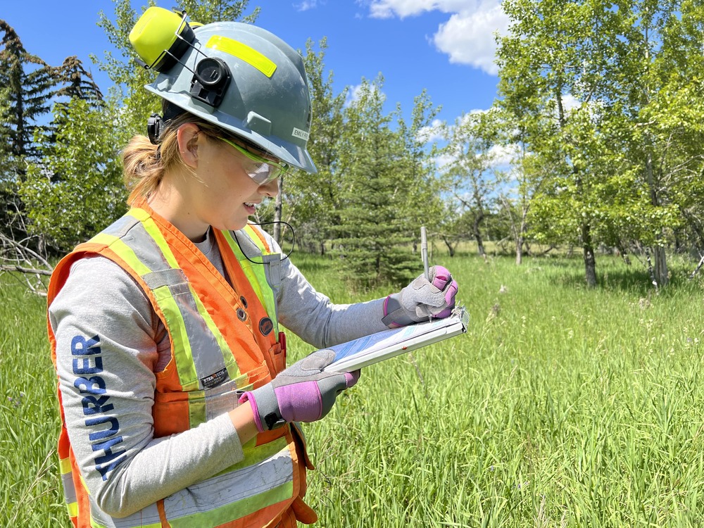 a woman in a helmet and safety vest writing on a clipboard in a field
