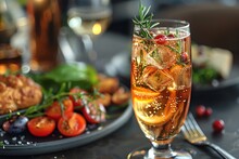 a glass of liquid with ice and a rosemary garnish next to a plate of food