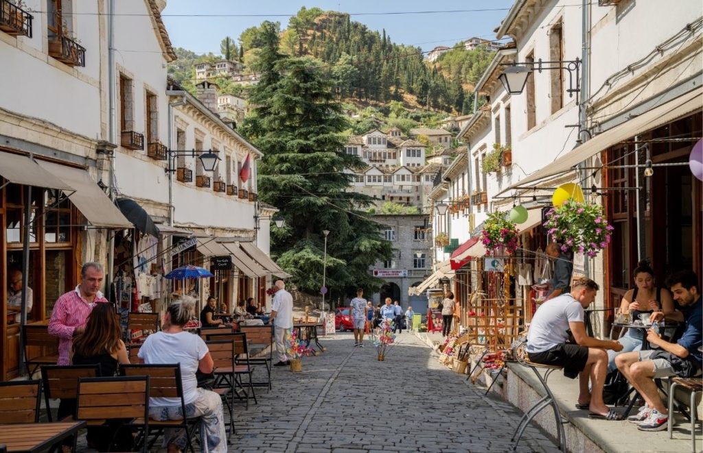 a street with people sitting on benches and tables in front of buildings with Plaka in the background