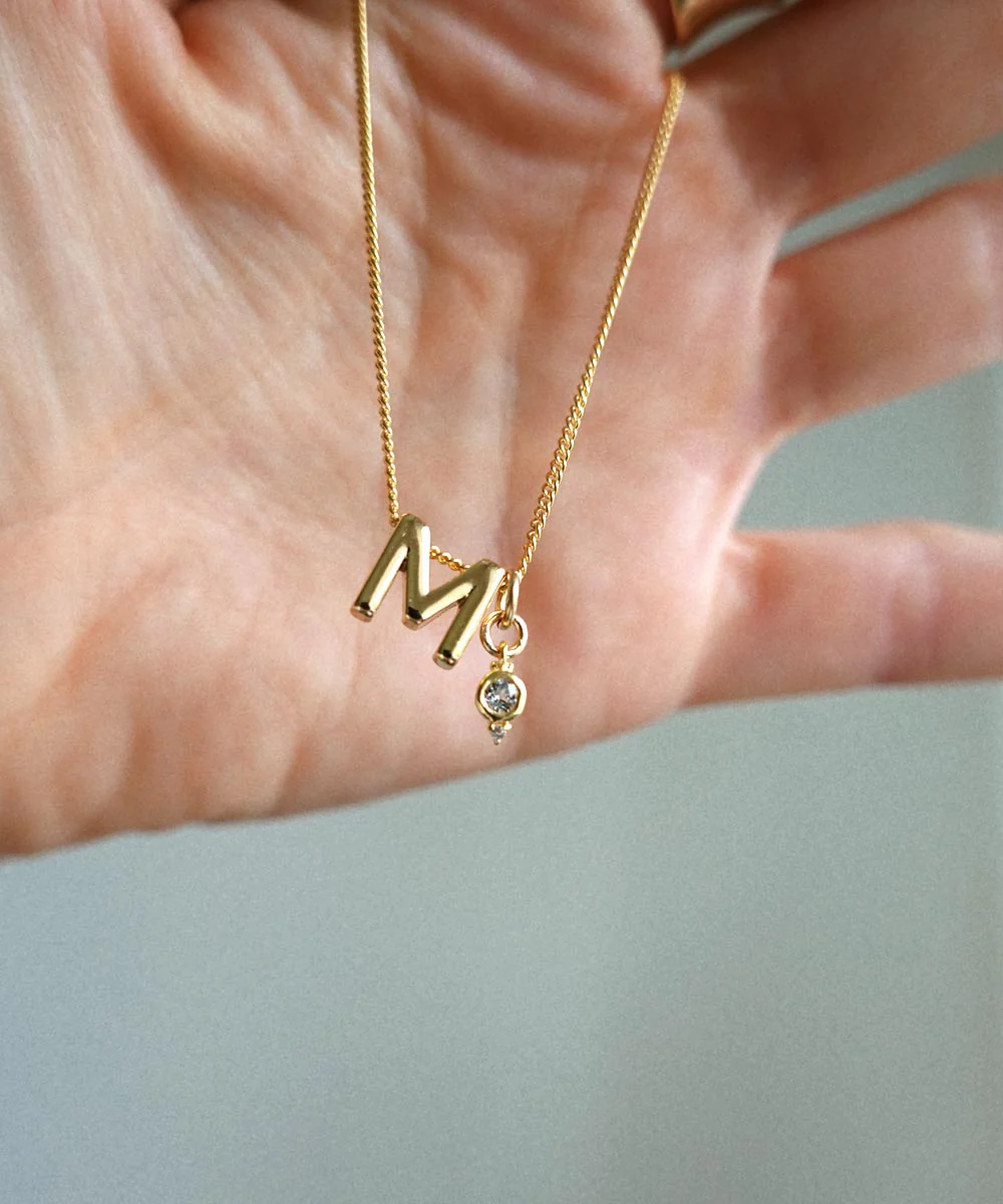 a hand holding a gold necklace with a letter on it
