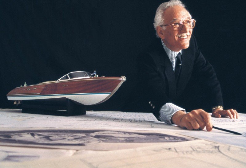a man in a suit and glasses sitting at a desk with a boat model