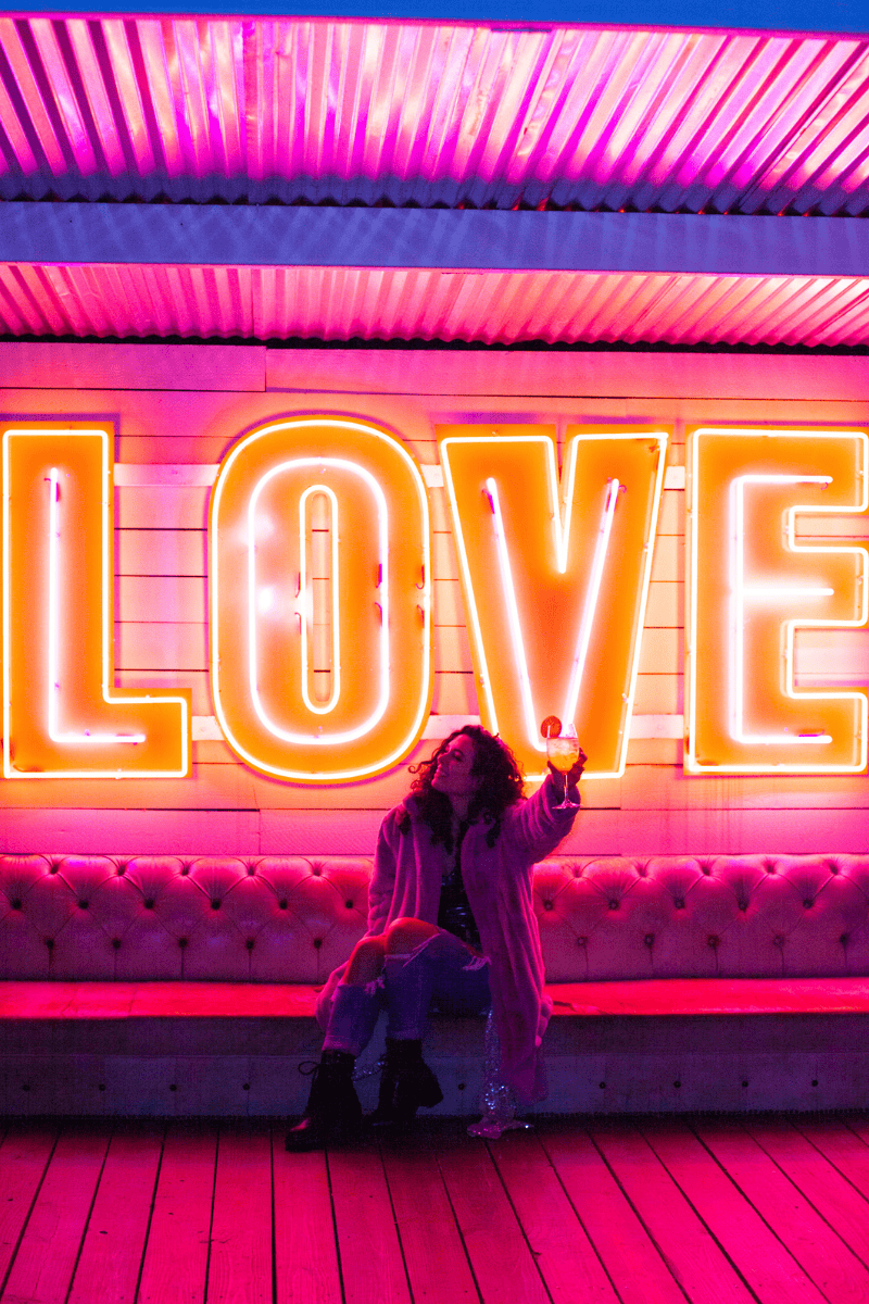 a woman sitting on a bench with a glass of wine in front of a neon sign