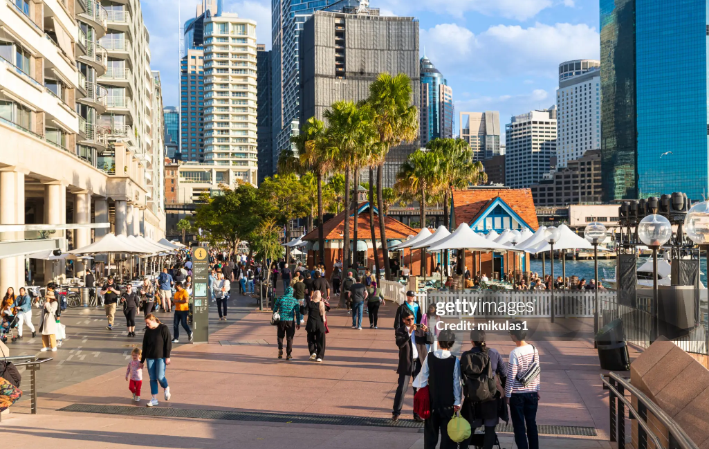 a group of people walking on a sidewalk with palm trees and buildings in the background