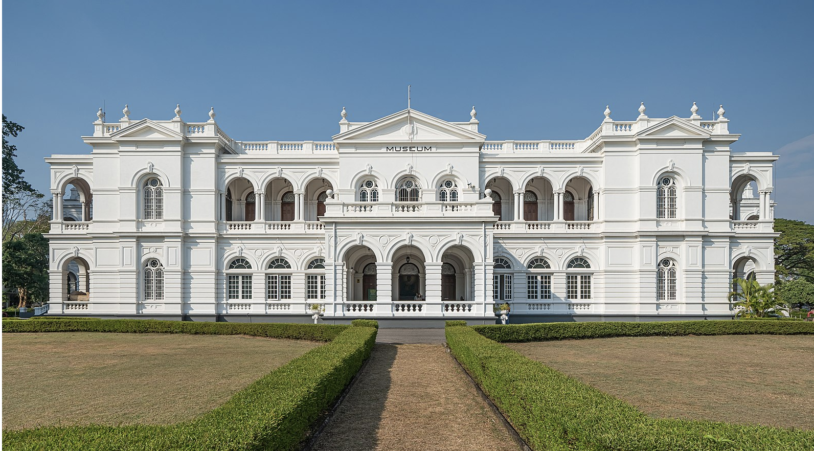 a white building with a path leading to the front with National Museum of Colombo in the background
