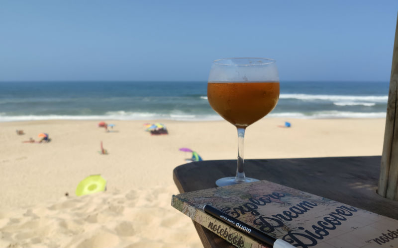 a glass of liquid on a table with a book and pen on a beach