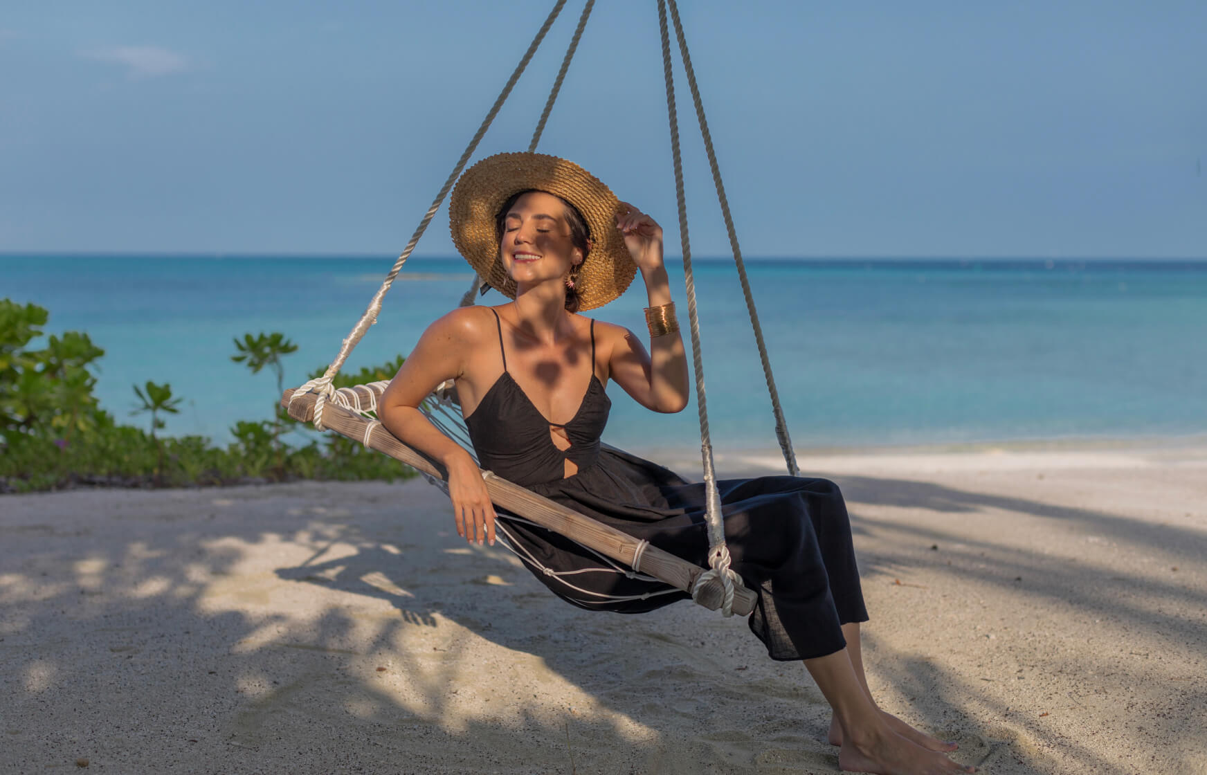 a woman in a black dress and hat sitting in a hammock on a beach