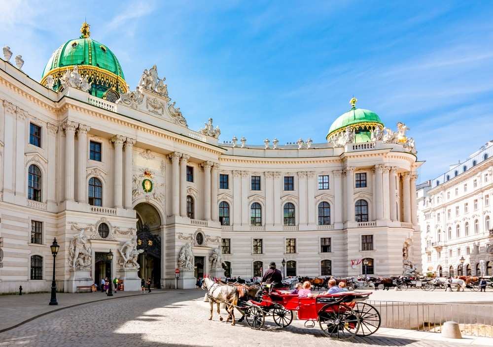 a horse carriage in front of a large white building with Hofburg in the background